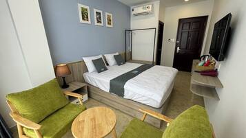 A hotel room with a bed, green upholstered chairs, a small wooden table, a TV mounted on the wall, and a door.