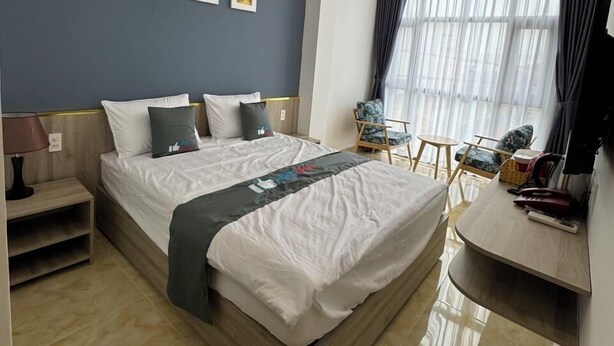 Une chambre d’hôtel moderne avec un grand lit, un bureau et une chaise. Trois tableaux encadrés ornent le mur, il y a une fenêtre avec des rideaux et un luminaire au plafond.