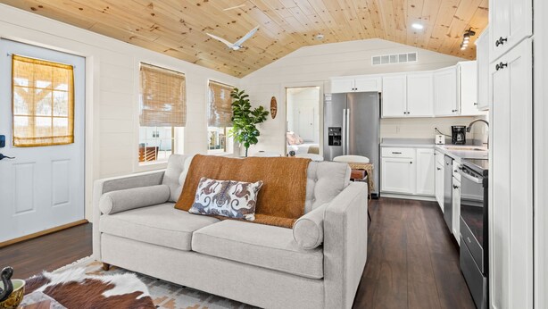 A cozy living room with a sofa, a coffee table, and a plant. The kitchen area includes white cabinets, a refrigerator, and a stove.