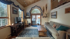 A cozy living room with a wooden ceiling, a television, a sofa, a coffee table, and a patterned rug. - Spectacular kid friendly home 2 minutes to Luzerne beach, 10min to Lake George! (Lake Luzerne)