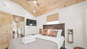 A bedroom with a wooden ceiling, a bed with a white and patterned bedspread, a wooden headboard, and a small shelf with a plant.