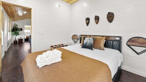 A bedroom with a wooden ceiling, a bed with a brown blanket, a white wall with three masks, and a wooden headboard.