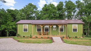A green single-story house with a porch, two windows on the front, and a gravel driveway.