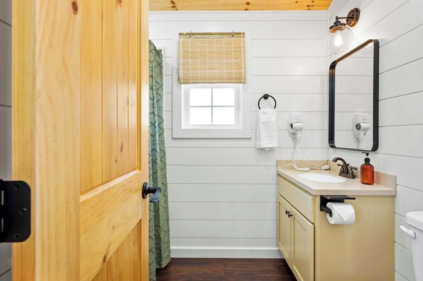 Un baño con puerta de madera, una ventana con persiana de bambú, una toalla colgada de un gancho, un lavabo con espejo y un inodoro.