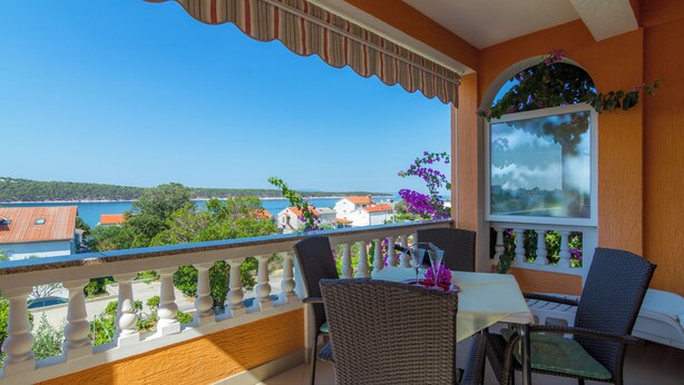 Un balcon agrémenté de meubles en osier, d’une table et offrant une vue sur la mer et les bâtiments.