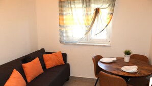 A room with a dark brown sofa, orange cushions, a wooden dining table with chairs, a potted plant, and a window with curtains. - Apartment mit Meerblick (Barbat)