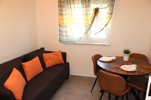 Ein Zimmer mit einem dunkelbraunen Sofa, orangefarbenen Kissen, einem Holzesstisch mit Stühlen, einer Zimmerpflanze und einem Fenster mit Vorhängen.