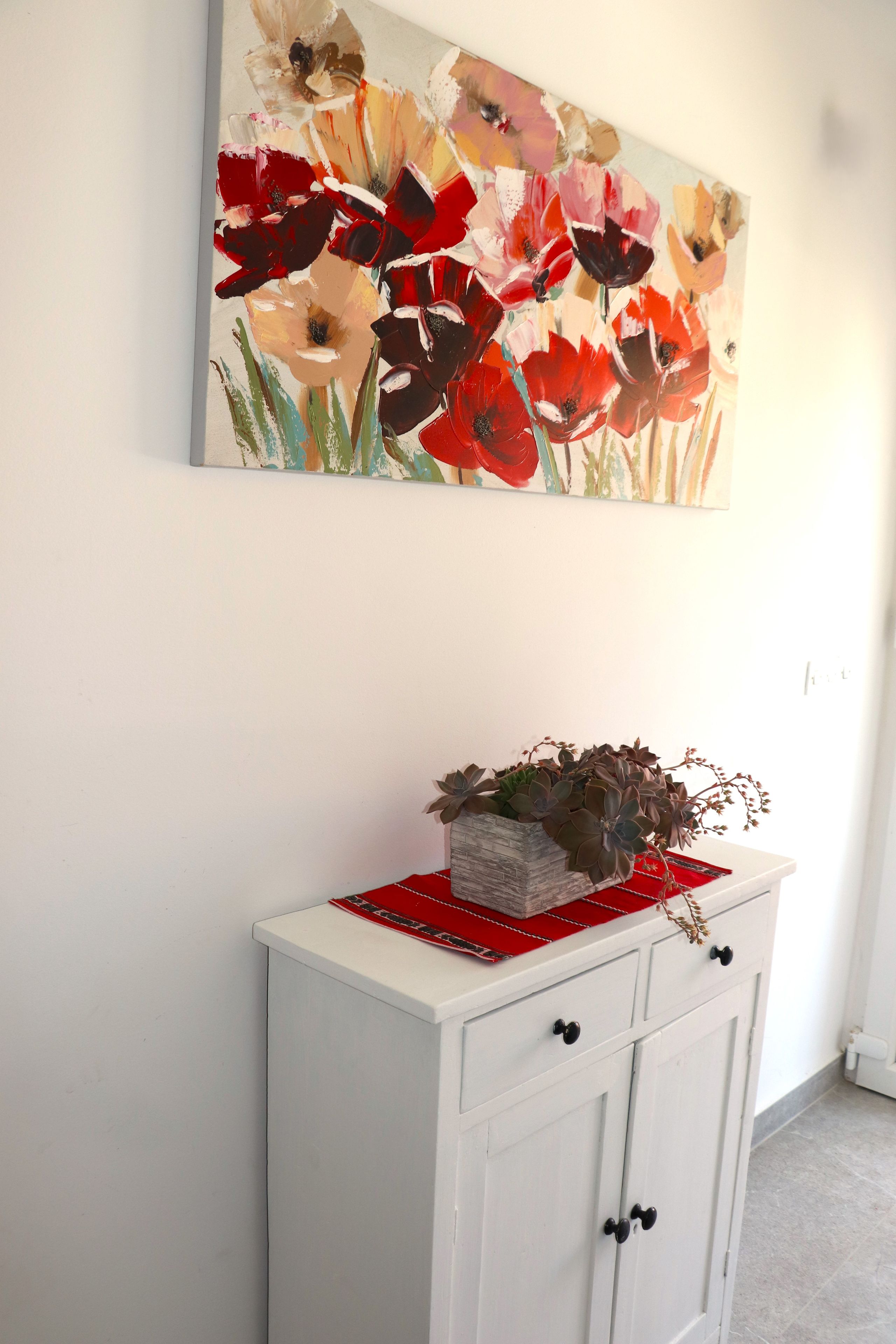 A white cabinet with black knobs, a red tray, and a floral painting above it.
