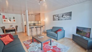 A modern living room with a sofa, a coffee table, a TV, a dining area with a table and chairs, and a kitchen in the background. - Apartment Hirle (Zermatt)