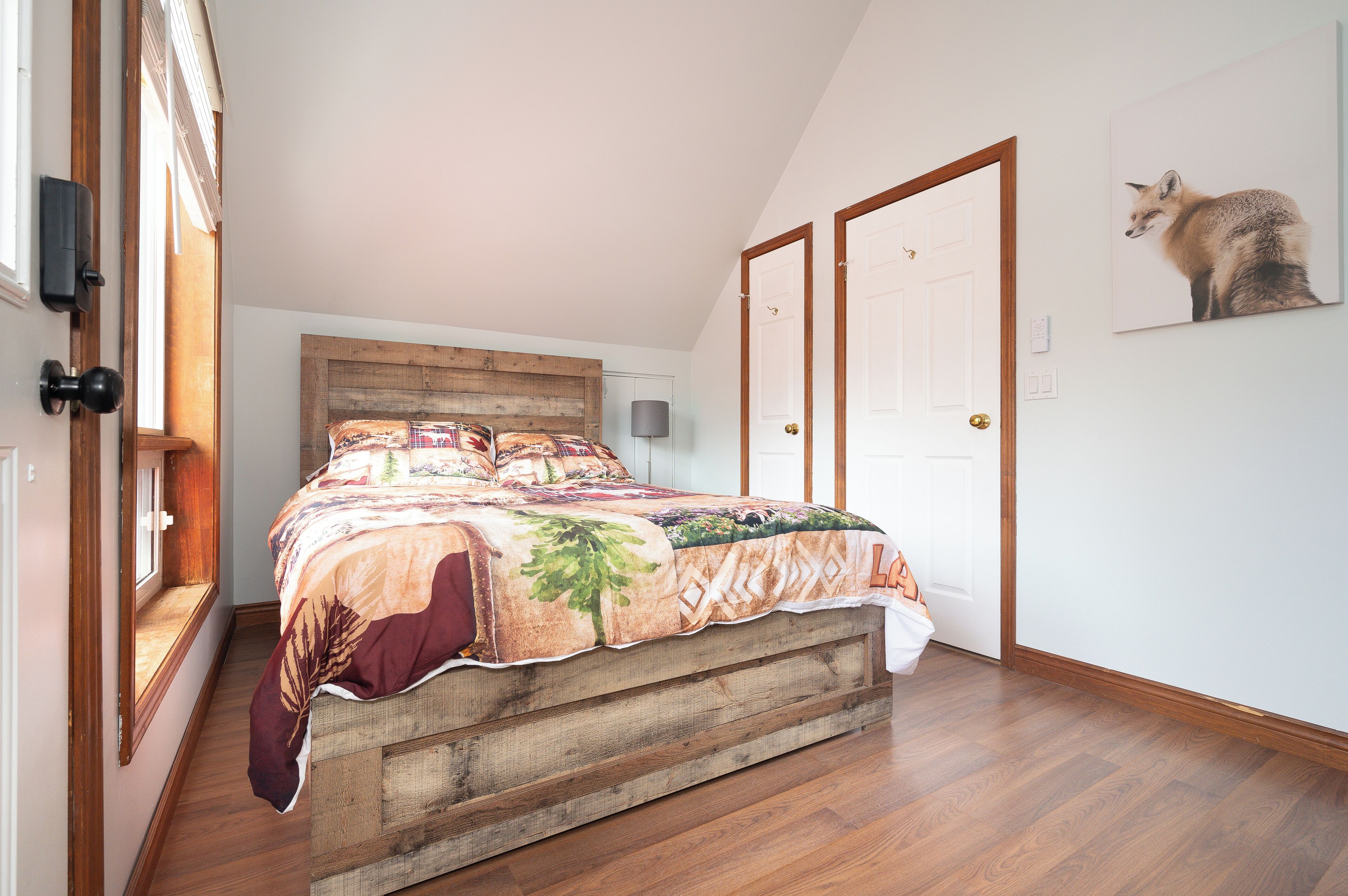 A bedroom with a wooden headboard, a large bed with a floral bedspread, a window with a view, and a fox painting on the wall.