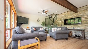 A modern living room with grey sofas, a wooden bench, a ceiling fan, a stone wall, and a staircase. - Cozy Family-Sized Cottage in Kearney - Hot Tub & Sauna 3 Min walk to Perry Lake (Kearney)