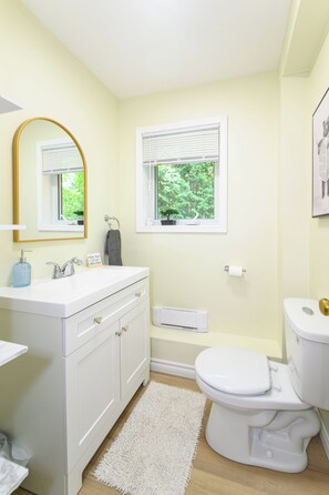 A bathroom with a white vanity, a gold-framed mirror, a window with blinds, a white toilet, and a white rug.
