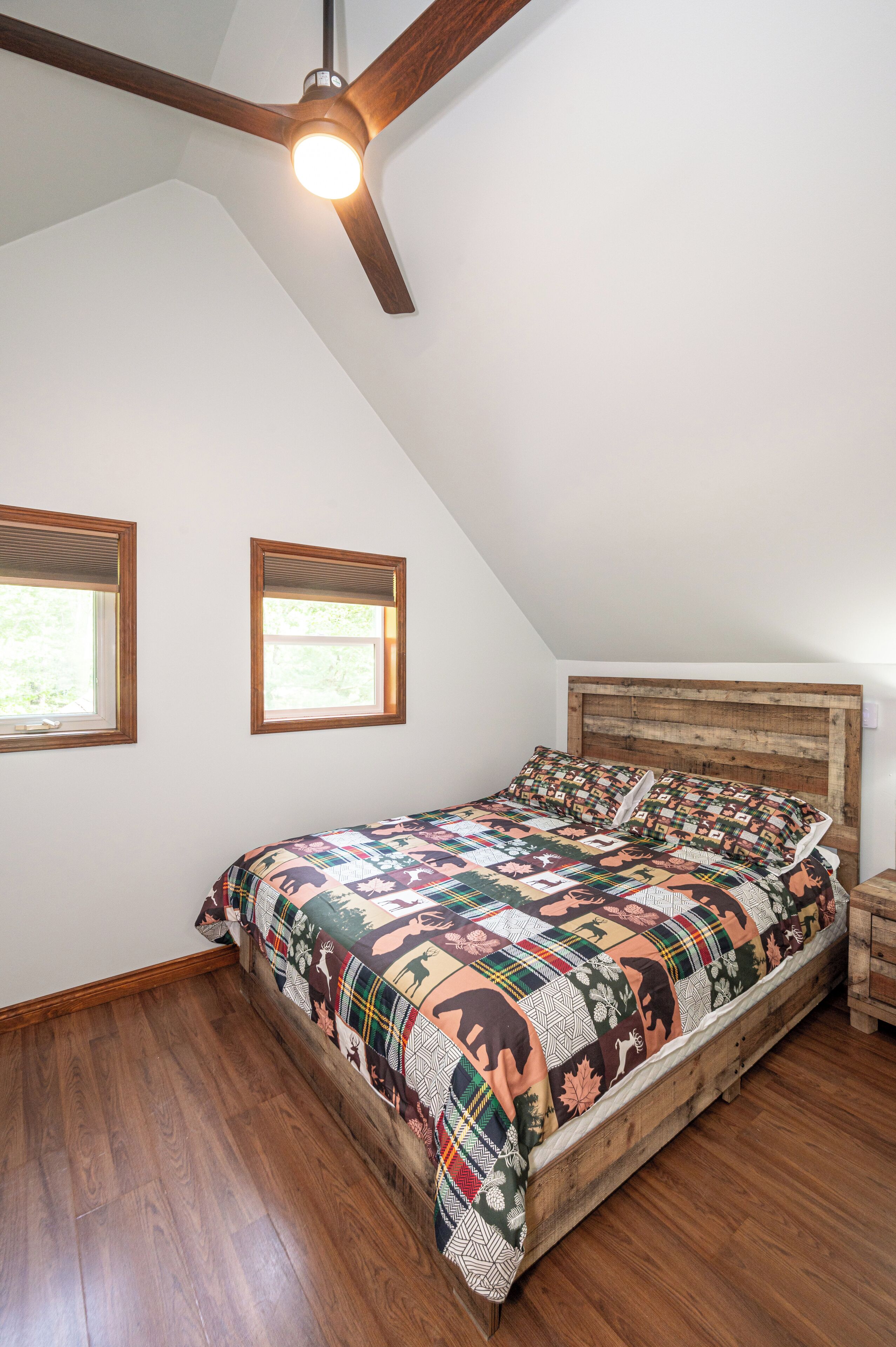 Une chambre avec un ventilateur de plafond en bois, deux fenêtres équipées de stores, un lit recouvert d’une courtepointe colorée et une tête de lit en bois.