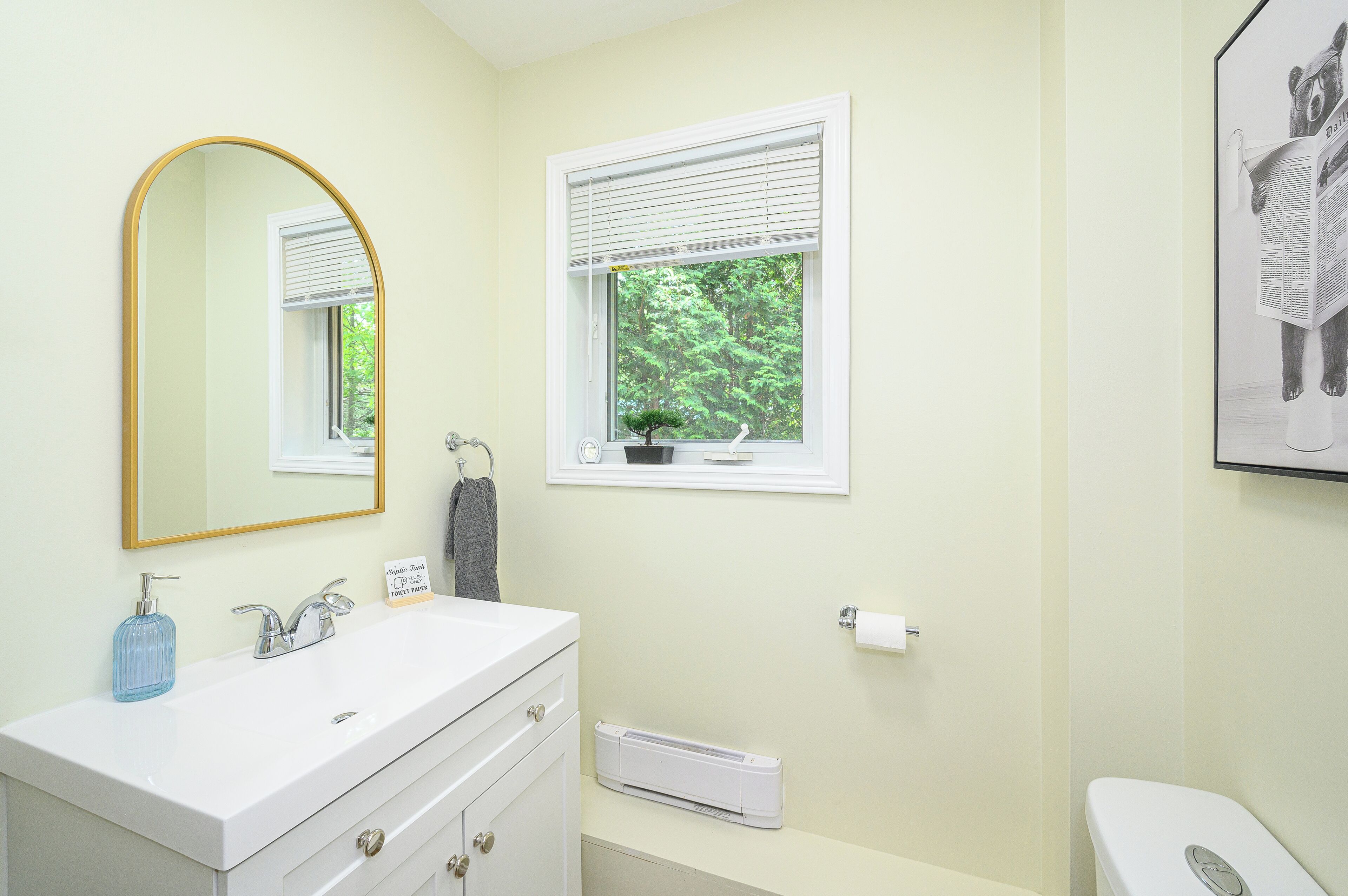 Une salle de bain avec un meuble-lavabo blanc, un miroir avec un cadre doré, une fenêtre avec des stores, des toilettes et un tableau encadré au mur.
