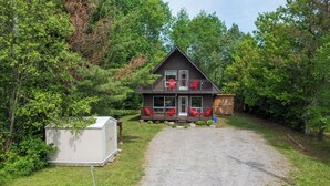 A two-story cabin with a balcony, a gravel driveway, and a shed in a wooded area. - Cozy Family-Sized Cottage in Kearney - Hot Tub & Sauna 3 Min walk to Perry Lake (Kearney)