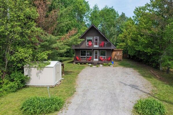 A two-story cabin with a balcony, a gravel driveway, and a shed in a wooded area.