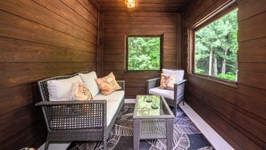 A wooden cabin interior with a sofa, two chairs, a small table, and a window providing a view of the forest. - Cozy Family-Sized Cottage in Kearney - Hot Tub & Sauna 3 Min walk to Perry Lake (Kearney)