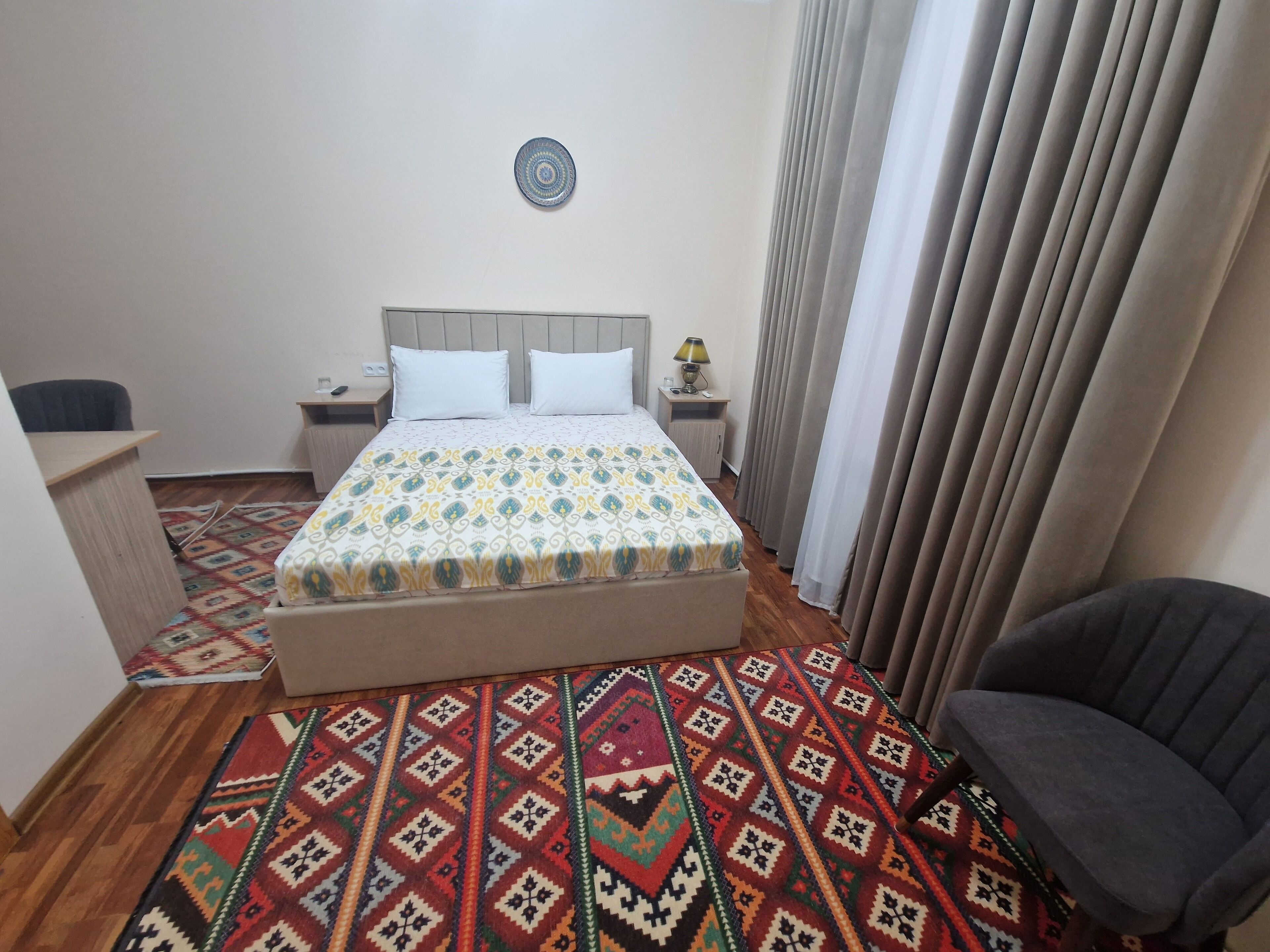 Quarto de hotel com cama, escrivaninha, cadeira, mesa de cabeceira e tapete estampado.