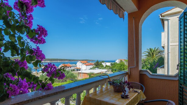 A balcony with a table and chairs, overlooking a coastal area with houses and a clear blue sky. - Apartment mit Meerblick (Barbat)