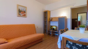A living room with an orange sofa, a television, a dining table with chairs, and a bookshelf. - Apartment mit Meerblick (Barbat)