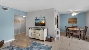 A living room with a TV, a dining table with chairs, a ceiling fan, and a wall-mounted sign that reads "BEACH HOUSE". - ☀️Steps2Beach-Pool View-Balcony-1BR-Villas on the Gulf G7☀️ (Pensacola Beach)