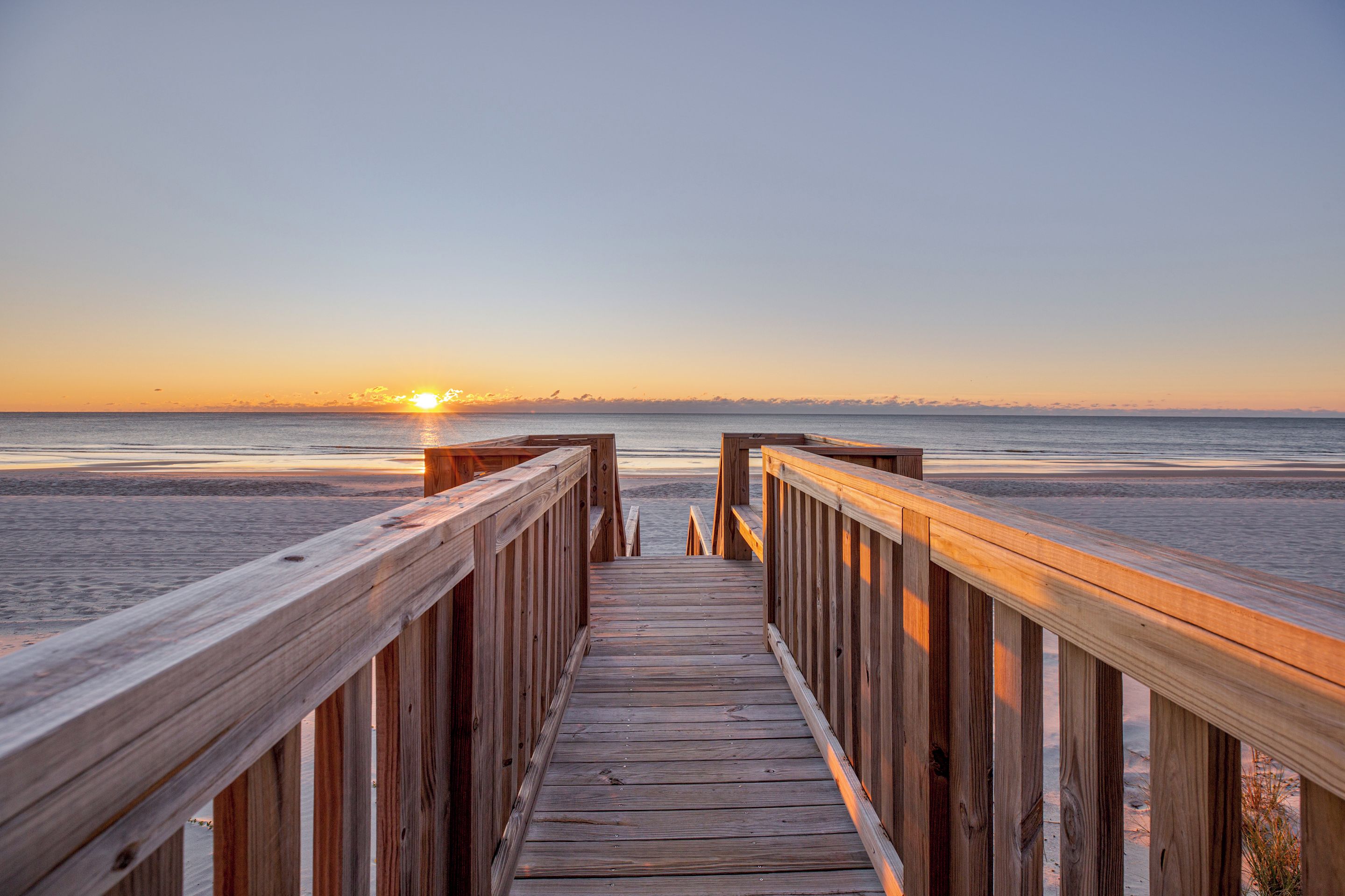 A wooden boardwalk leading to a beach with the sun setting in the distance.