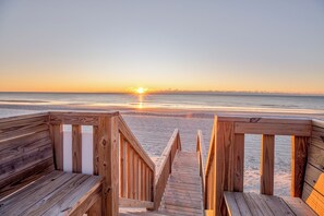 Ein hölzerner Steg führt zu einem Strand, im Hintergrund ist ein Sonnenuntergang zu sehen.