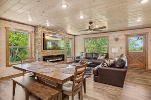 A spacious living area with a fireplace, wooden furniture, and large windows offering a view of greenery. - Hilltop Dreamin' 4 Bedroom Cabin by RedAwning (Sevierville)