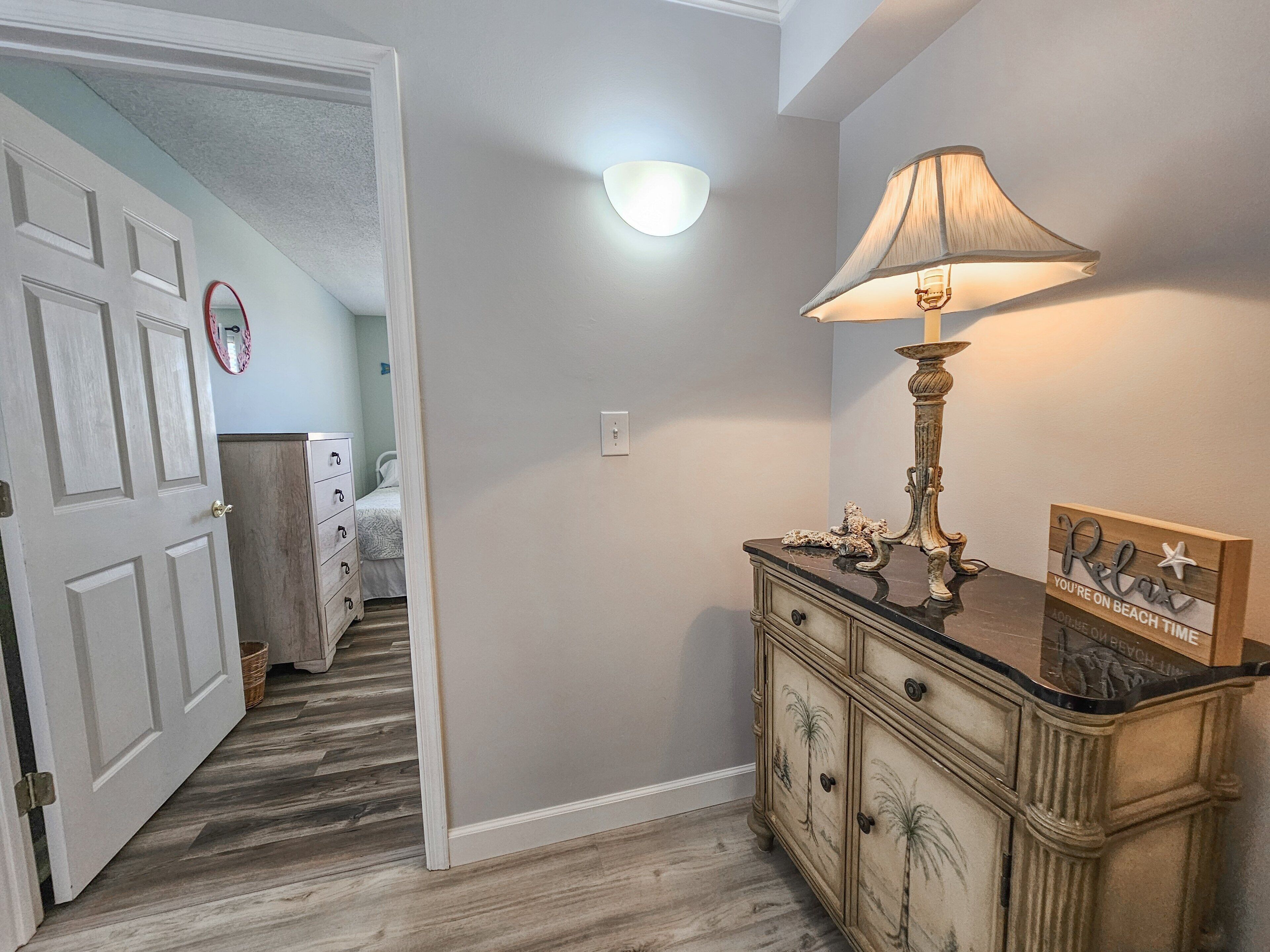 A hallway with a wooden floor, a white door, a dresser with a lamp, and a wall with a light fixture.