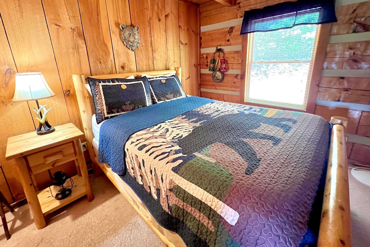 A wooden cabin bedroom with a bed, a nightstand, a lamp, and a window with a view.