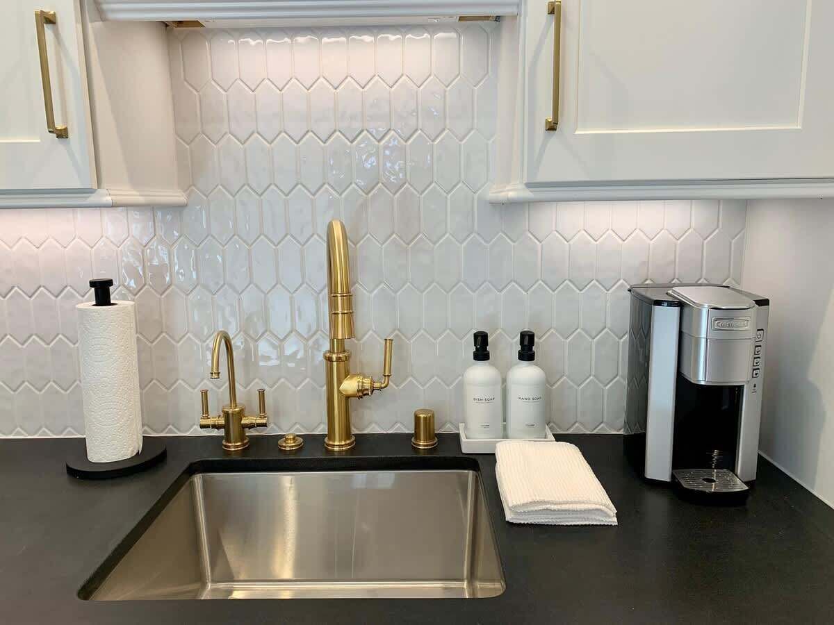 Eine Küche mit Spüle aus Edelstahl, einem goldenen Wasserhahn, einer Kaffeemaschine und Toilettenartikeln auf der Arbeitsfläche.