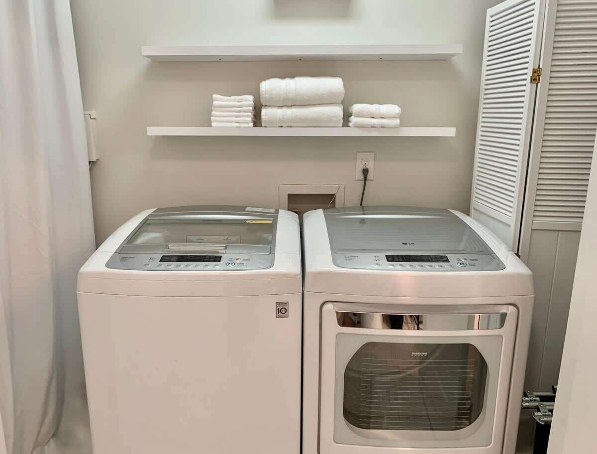 A laundry area with two front-loading washers and a dryer, a white shelf with folded towels, and a door with a louvered design.