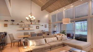 A spacious living room with a large sectional sofa, a wooden coffee table, and a chandelier. There are multiple shelves with decorative items and a dining area with a table and chairs.