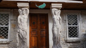 Ein Steinhaus mit einer Holztür, zwei Statuen an jeder Seite und einem kleinen Balkon mit Metallgitter.