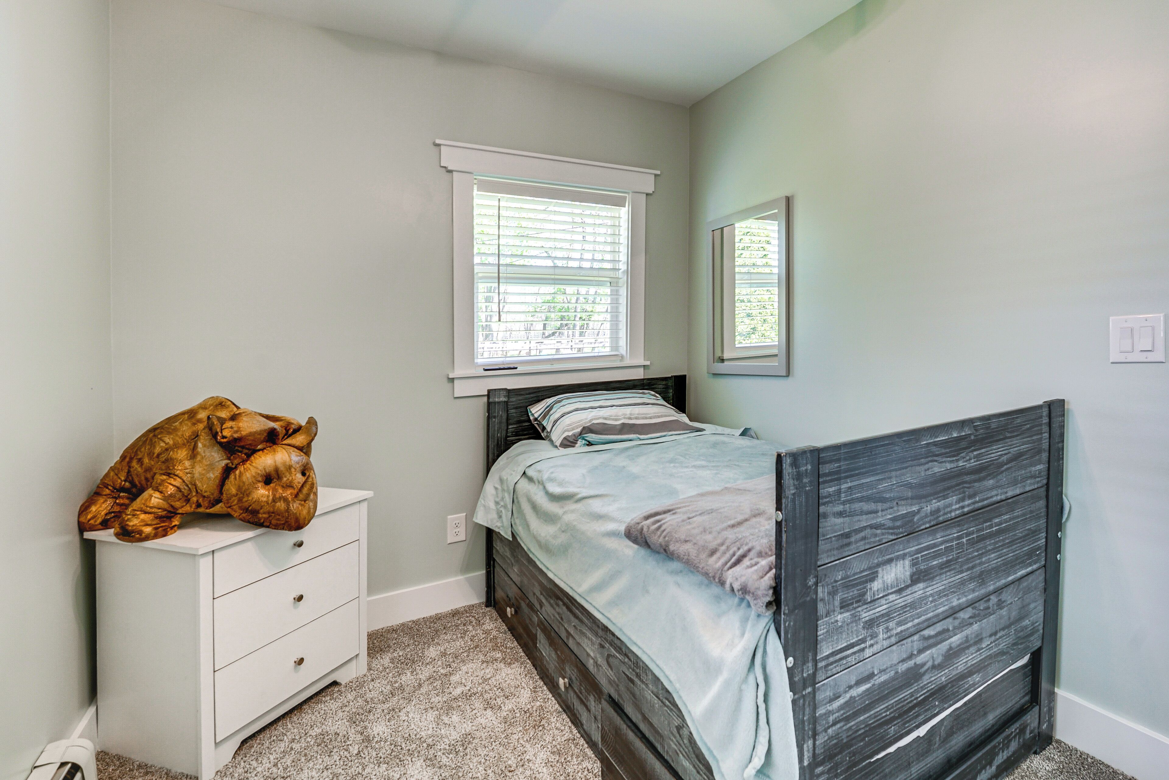 A bedroom with a bed, a nightstand, a window with blinds, and a mirror on the wall.