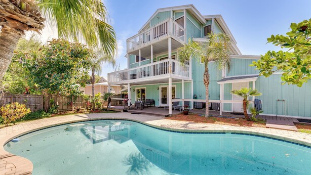 A two-story house with a swimming pool, surrounded by palm trees and a wooden deck.