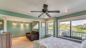 A bedroom with a bed, a ceiling fan, a desk, a chair, and a balcony with a view of greenery.