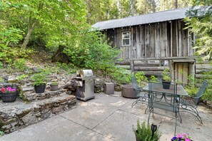 Une cabane en bois rustique, avec une table et des chaises en métal, entourée de plantes en pot et d’un barbecue.