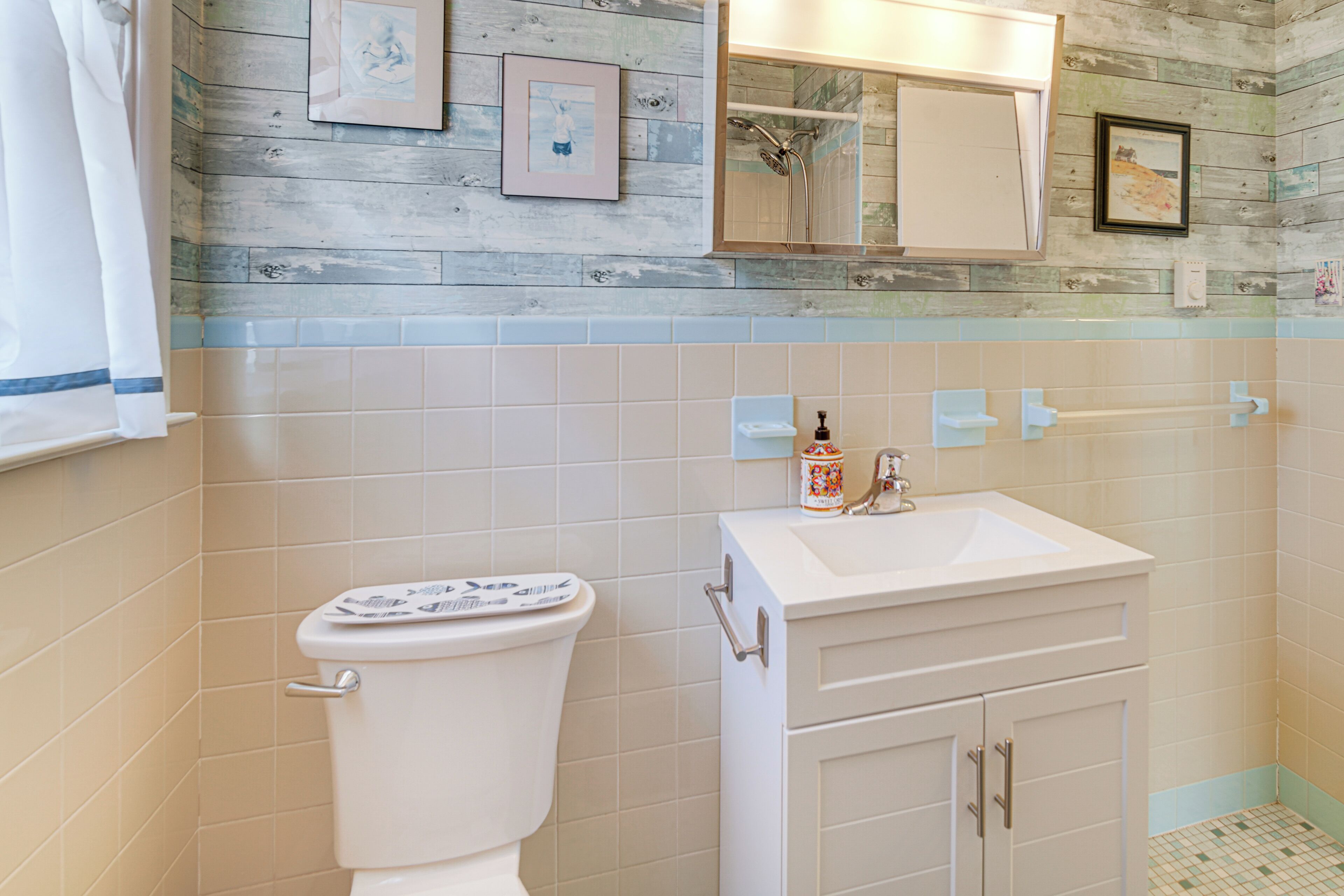 Een badkamer met toilet, wastafel en spiegel. Er hangen afbeeldingen in lijsten aan de muur en is er een douchehoek.