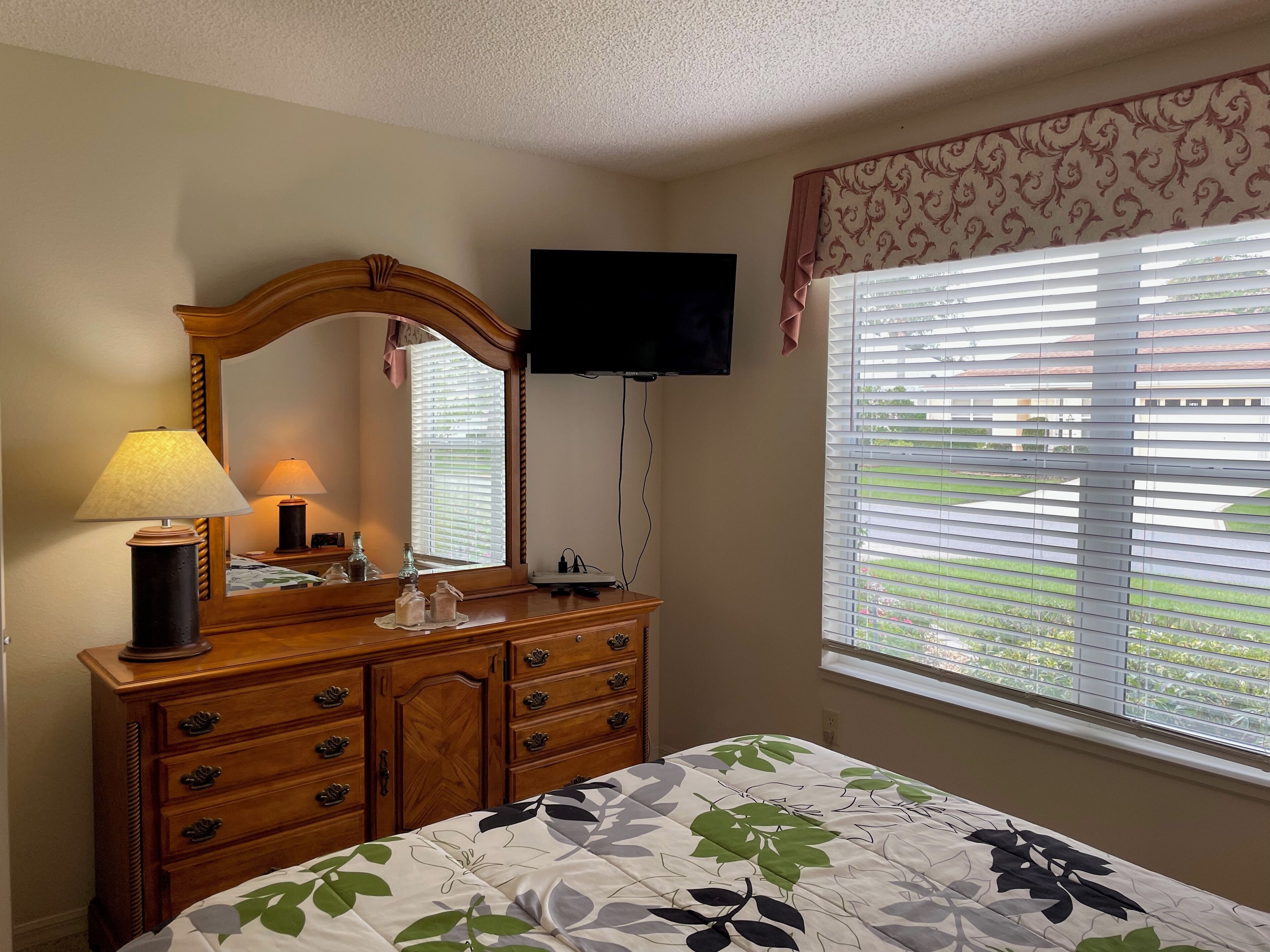 A bedroom with a bed, dresser, mirror, lamp, and a TV on the wall.