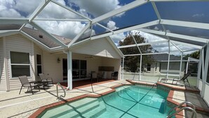 A covered outdoor swimming pool with a patio area, chairs, and a view of a house in the background. - Private Pool Paradise close to Lake Sumter Landing Market Square! (The Villages)