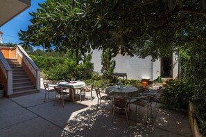 An outdoor patio with a round table and chairs, surrounded by greenery and a white building in the background. - Behindertengerechte Wohnung im Erdgeschoss (Barbat)