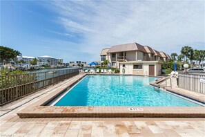 A swimming pool with a wooden deck, a lifeguard stand, and a building with a brown roof in the background. - Good Times & Tan Lines (Fort Myers Beach)