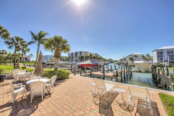 A waterfront patio with outdoor seating, a marina with boats, and modern buildings in the background. - Good Times & Tan Lines (Fort Myers Beach)