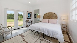 A bedroom with a large bed, a wooden headboard, a white rug, and a view through a glass door.