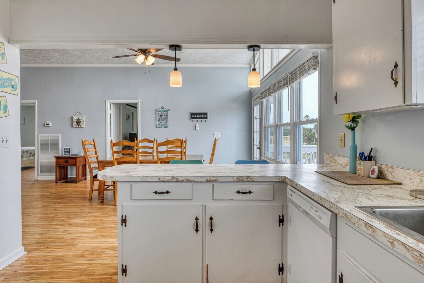 Une cuisine avec des meubles blancs, une table à manger en bois et des chaises. On y trouve un ventilateur de plafond, des luminaires suspendus et une vue dégagée grâce à de grandes fenêtres.