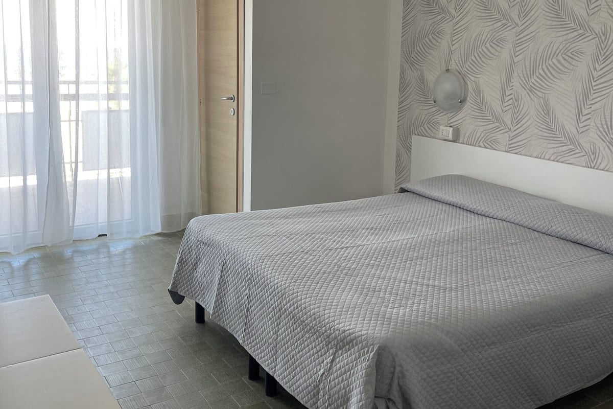 A bedroom with a bed, a door, a window with sheer curtains, and a patterned wall.