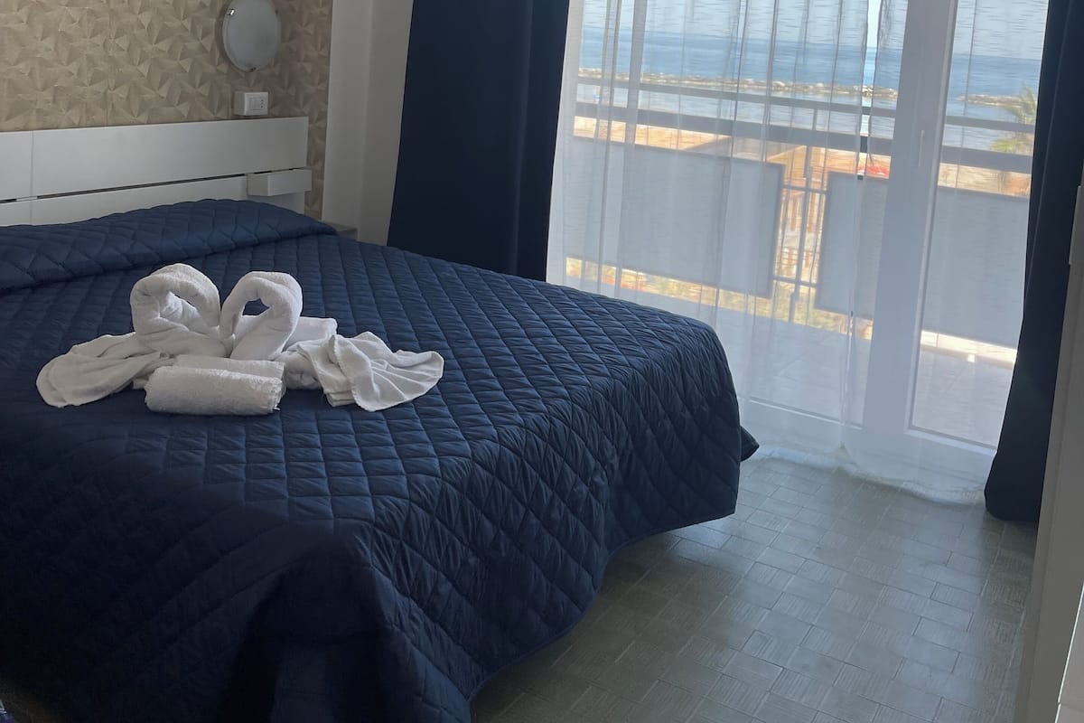 A hotel room with a bed, a wall-mounted toilet, a window with curtains, and a view of the sea.