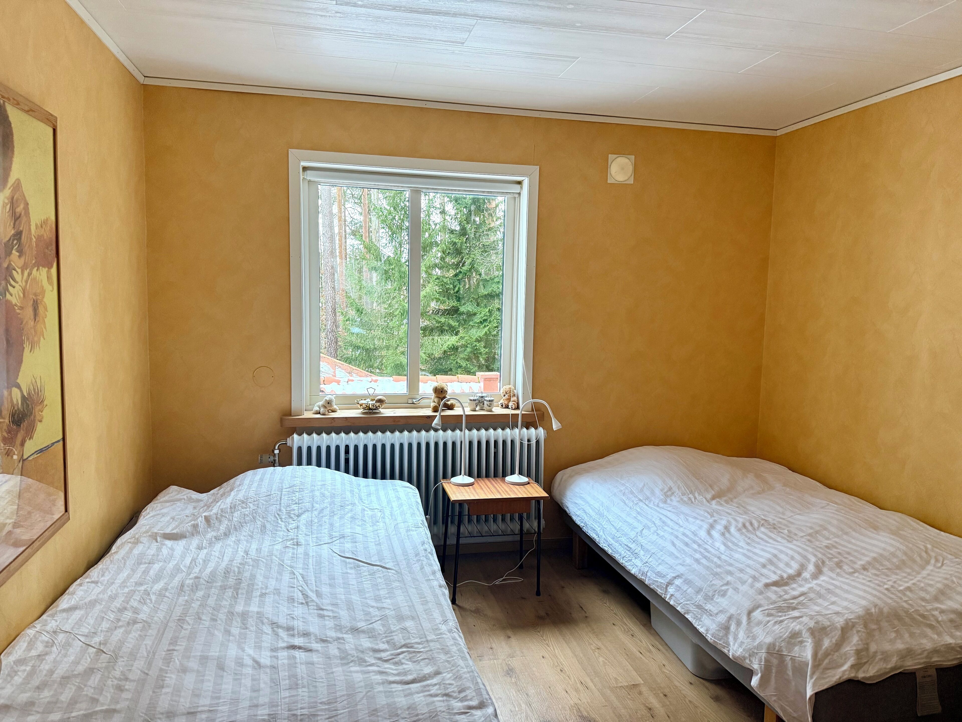 A room with two single beds, a radiator, a small table, and a window with a view of trees.