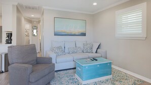 A living room with a grey armchair, a light grey sofa with patterned cushions, a blue storage chest, and a painting of sailboats on the wall.